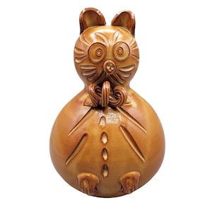 May include: A ceramic figurine of a stylized cat with large eyes, ears, and a bow tie. The figurine is a warm brown color with carved details, including lines on the face and body. The cat has a rounded body and small feet.