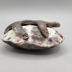 May include: A small ceramic sculpture of a brown and white animal curled up on a pillow. The animal has a long, thin body and small, rounded ears. The pillow is white with brown and red accents.
