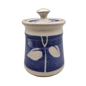 May include: A white ceramic jar with a blue and white design. The jar has a lid with a knob on top. The design features stylized leaves in white on a blue background.