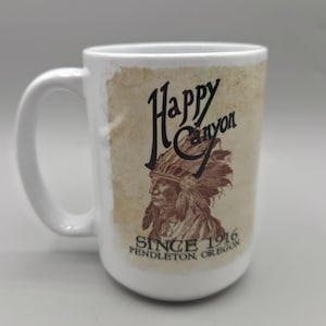 May include: White ceramic mug with a large handle. The mug features the text "Happy Canyon" in a stylized font, along with an illustration of a Native American chief. The text "SINCE 1916 PENDLETON, OREGON" is also included.