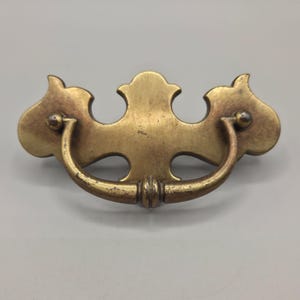 May include: A brass-toned metal drawer pull featuring a decorative backplate with curved, symmetrical edges and a central crest. A U-shaped swinging handle is attached to the backplate by two small circular mounts, showing a weathered finish.