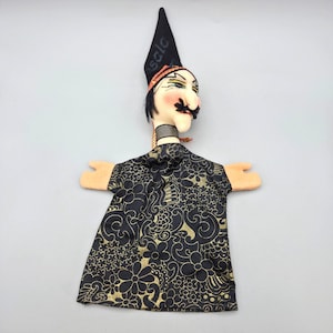 May include: A hand puppet featuring a character with a pale face, black mustache, and dark hair. The puppet wears a tall, pointed black hat with faint text and a black and gold floral patterned tunic. The hands are light peach-colored fabric.