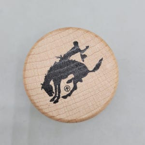 May include: A round, wooden coaster with a black silhouette of a bucking bronco and rider. The coaster has a light wood grain pattern and a simple, rustic design. The image is centered on a light gray background.