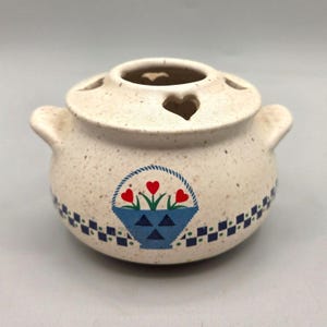 May include: A speckled ceramic pot with a lid featuring heart-shaped cutouts. The pot has two handles and a blue basket design with red heart-shaped flowers. A blue and white checkered pattern decorates the base.