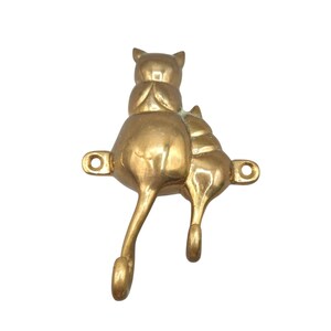May include: A polished brass wall hook featuring two stylized cats sitting side by side. The tails of the cats extend downward to form two separate hooks for hanging items. The piece includes two horizontal mounting tabs with circular screw holes.