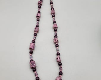 May include: A long necklace made of dark purple and light pink beads. The beads are cylindrical and have a decorative pattern. The necklace is strung on a gold chain.