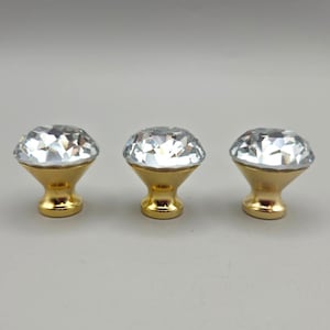 May include: Three cabinet knobs featuring a clear, faceted, diamond-shaped glass top set into a polished gold-tone metal base. Each knob has a wide, circular crystal head that tapers down to a smooth, rounded gold pedestal for attachment.