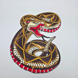 May include: An embroidered patch featuring a coiled rattlesnake with its mouth open, displaying sharp fangs and a long red tongue. The snake has brown, tan, and black scales with a distinct red and white patterned border along its underside.