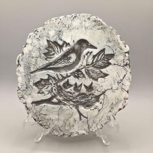 May include: A round, off-white ceramic plaque with a bird and nest relief. The bird is perched on a leafy branch above a nest with three baby birds. The plaque has a cracked, textured surface and is displayed on a clear stand.