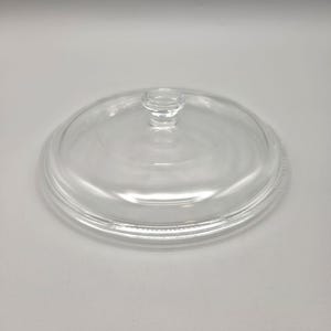May include: A circular, transparent glass lid featuring a smooth, slightly domed surface and a centered, raised knob handle. The lid has a distinct, stepped rim design around the outer edge, providing a secure fit for a corresponding container.