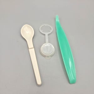 May include: A set of three plastic items including a cream-colored spoon, a small white circular component with a thin stem and ring top, and a long, curved mint green tool with a pointed end, all arranged side by side on a neutral surface.