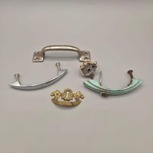 May include: A collection of four assorted metal drawer pulls and hardware pieces. Included are a silver-toned handle with mounting plates, a curved chrome pull, a small brass-colored keyhole escutcheon, and a distressed light blue curved handle.