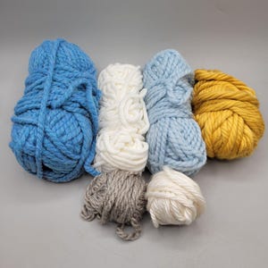 May include: A collection of colorful yarn skeins in various shades of blue, white, gray, and yellow. The yarn appears to be thick and chunky, suitable for knitting or crocheting projects. The arrangement of the yarn balls creates a visually appealing display.