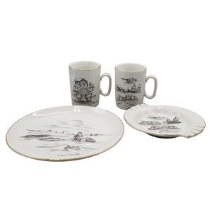May include: A set of white ceramic mugs and plates featuring black and gold illustrations of people and dogs in a snowy landscape. The mugs have a gold rim and the plates have a gold trim.