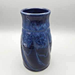 May include: A cylindrical ceramic vase with a glossy finish, featuring a deep navy blue base color that transitions into a lighter, mottled cobalt blue glaze near the rim, with subtle vertical swirling patterns throughout the body.