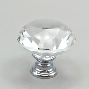 May include: A clear, faceted, diamond-shaped cabinet knob with a polished chrome base. The top portion is transparent and cut with multiple geometric angles to reflect light, while the circular metal base provides a sturdy mounting point.