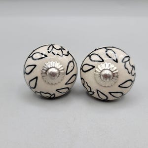 May include: Two cream-colored ceramic cabinet knobs with black leaf-like patterns. Each knob features a silver-colored, flower-shaped center. These decorative knobs are ideal for adding a unique touch to furniture or cabinets.