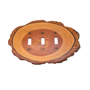 May include: A triple light switch cover plate crafted from a natural wood slice with a visible grain pattern and rough, textured bark edges. The oval-shaped wooden plate features three rectangular cutouts for switches and four small screw holes.