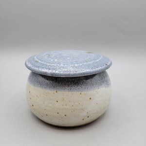 May include: A handmade ceramic butter dish with a lid. The dish is round with a cream-colored base and a gray-blue top. The lid is also gray-blue. The dish has a rustic, speckled appearance.