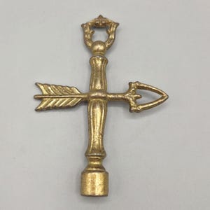 May include: A gold-colored metal bottle stopper with an arrow and a decorative handle. The arrow is horizontal, and the handle has a loop at the top. The stopper has a vintage or antique appearance.