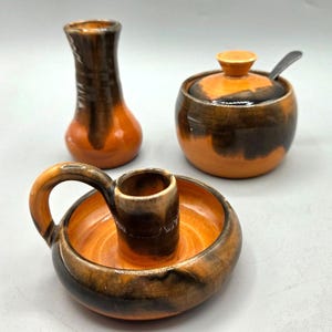 May include: A ceramic set featuring a small vase, a sugar bowl with a spoon, and a candle holder. The pieces are glazed in shades of orange and brown, with a glossy finish. The candle holder has a handle.