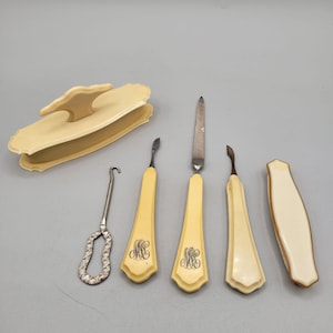 May include: A collection of antique ivory-colored manicure tools, including a nail file, cuticle pushers, and a nail buffer. The handles of the tools are engraved with the initials "MC". The set also includes a small silver tool and a matching ivory-colored case.