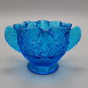 May include: A blue glass bowl with a ruffled rim and two wing-shaped handles. The bowl has a diamond pattern and is on a pedestal base.