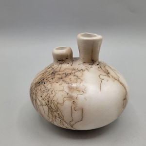 May include: A handcrafted, off-white ceramic vase with two openings. The vase has a rounded body with a textured, crackle-like pattern in brown and black. The openings are cylindrical and smooth.