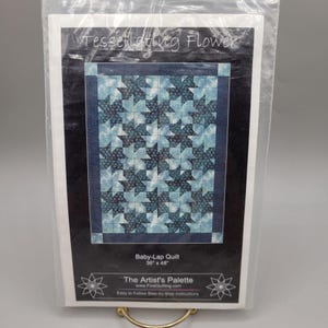 May include: A packaged quilt pattern titled "Tessellating Flower." The pattern features a baby-lap quilt design with a blue and white star pattern. The quilt measures 91 cm x 122 cm and is from The Artist's Palette.