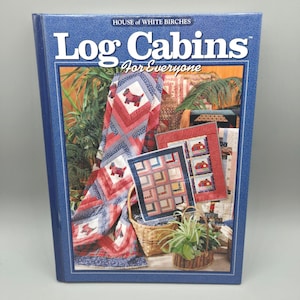 May include: A blue book titled "Log Cabins For Everyone" by House of White Birches. The cover features a quilt with a red and blue pattern and a dog design. Other quilts and a basket are also visible.