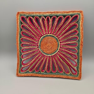 May include: A square woven trivet featuring a central orange circular disc surrounded by radiating pink petal-like loops. The design is framed by concentric borders in teal and orange, creating a vibrant, multi-colored floral pattern.