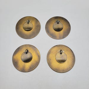 May include: Four brass-toned metal pendants. Each pendant has a large, circular base with a smaller, circular accent piece attached at the centre. The pendants have a vintage, aged appearance with gold and bronze hues.