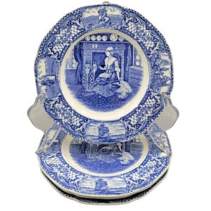 May include: A stack of blue and white decorative plates. Each plate features a central scene of a woman spinning wool, surrounded by a detailed border. The plates are displayed on a clear plastic stand, showcasing the intricate design.