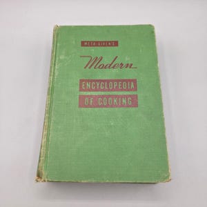 May include: A green hardcover book titled Meta Given's Modern Encyclopedia of Cooking. The front cover features dark red rectangular blocks with white text reading Meta Given's, Modern, Encyclopedia, and of Cooking in a clean font style.