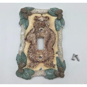 Peut inclure: Plaque décorative pour interrupteur avec un ours brun au centre. Le cadre est orné de feuilles vertes et de pommes de pin marron. Deux vis sont incluses. Conçu pour un interrupteur standard, il ajoute une touche rustique.