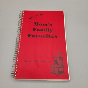 May include: A red spiral bound cookbook titled "Mom's Family Favorites" by Nellie Delia Vannice. The cover features a rolling pin and a wood-burning stove.