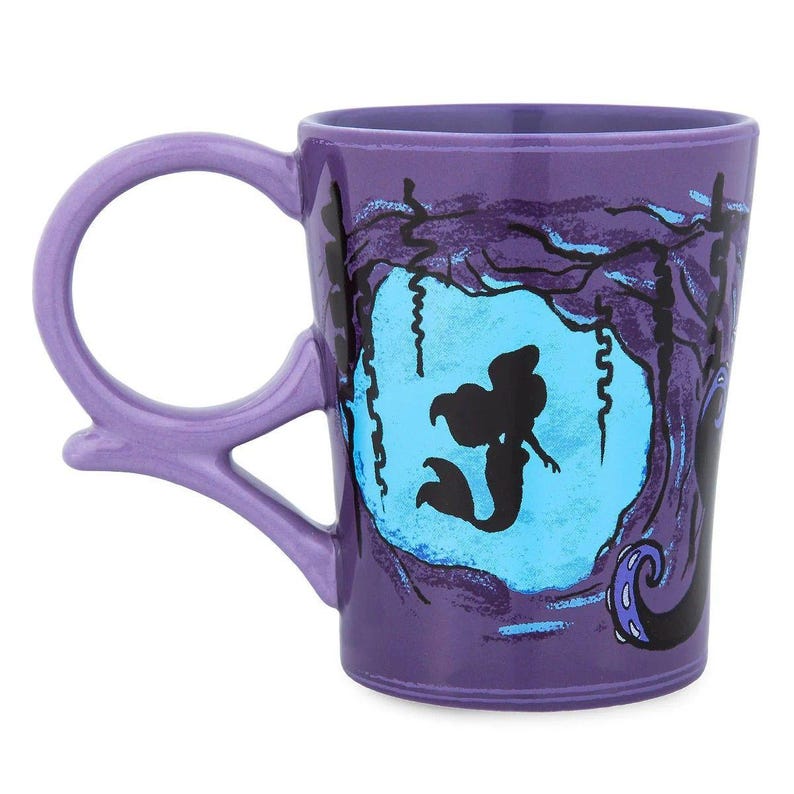 May include: A purple ceramic mug featuring a silhouette of a mermaid within a blue oval, surrounded by dark tree-like shapes. The mug has a unique handle design and is a collectible item.