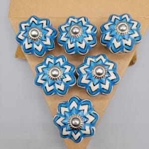 May include: Seven blue and white floral ceramic cabinet knobs with silver centres. The knobs are arranged on a brown triangular piece of cardboard against a grey background. Each knob has a unique flower design.