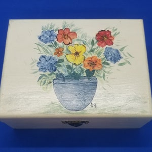 May include: A white wooden box with a painted design of a blue vase filled with yellow, red, and orange flowers. The box has a brass latch.