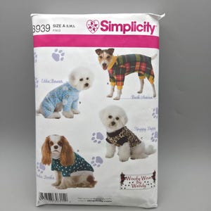 May include: A Simplicity sewing pattern envelope 3939 for dog clothing in sizes S, M, and L. The white package features illustrations of four dogs wearing various outfits, including a plaid jumpsuit, a blue patterned pajama, a leopard print shirt, and a heart-print top.