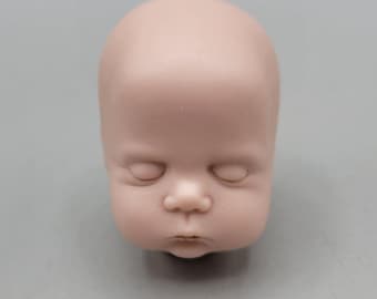 May include: A pale pink doll head with closed eyes, a small nose, and a closed mouth. The doll head is smooth and appears to be made of a solid material. The head is a realistic representation of a baby's face.