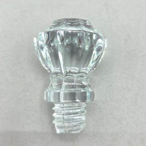 May include: Clear glass bottle stopper with a faceted, decorative top. The stopper has a threaded base for secure fitting. The design features a series of vertical ridges and a detailed, rounded top. The clear glass allows light to pass through.