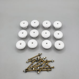 May include: A set of 10 white ceramic cabinet knobs with a single hole for installation. The knobs are arranged in a grid pattern. The set also includes 10 gold-colored screws for attaching the knobs to cabinets.
