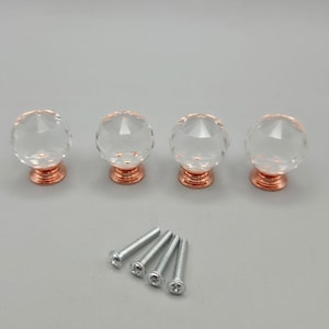 May include: Four clear, faceted, spherical glass knobs with metallic rose gold bases, arranged in a horizontal row. Below the knobs are four silver-colored metal machine screws with flat heads, intended for installation on furniture or cabinetry.
