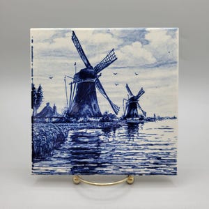 May include: Square ceramic tile featuring a blue and white illustration of two windmills on a body of water. The artwork depicts a rural landscape with trees, a thatched-roof building, and birds in the sky. The tile is displayed on a gold-colored stand.