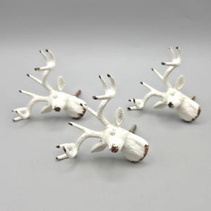 May include: Three white metal cabinet knobs shaped like deer heads with antlers. Each knob features a distressed finish with brown accents on the nose and antler tips, and a small metal screw extending from the back for mounting purposes.