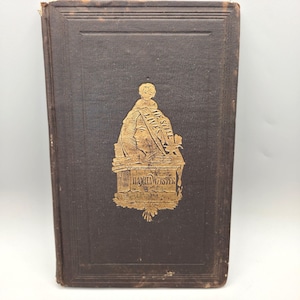 May include: A vintage hardcover book with a dark brown cover. A gold-colored emblem featuring a portrait and the text "Daniel Webster" is centered on the cover. The book's title is "He Still Lives".