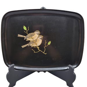 May include: Black rectangular tray with rounded corners, featuring a gold and brown bird design on a branch with green leaves. The tray is displayed on a black stand, suitable for serving or display.