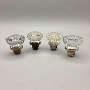 May include: Four clear glass cabinet knobs with a faceted design. Each knob has a metal base in varying colors, including gold and silver. The knobs are likely vintage or antique.