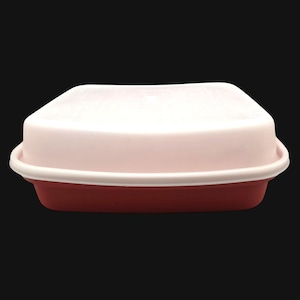 May include: A rectangular food container with a white lid and a red base. The lid is slightly curved and fits snugly on the base. The container has a white rim. The background is black.